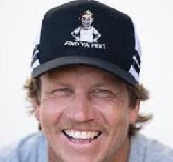 A photo of Tommy standing facing the camera with a hat on that has the words, Find ya feet
